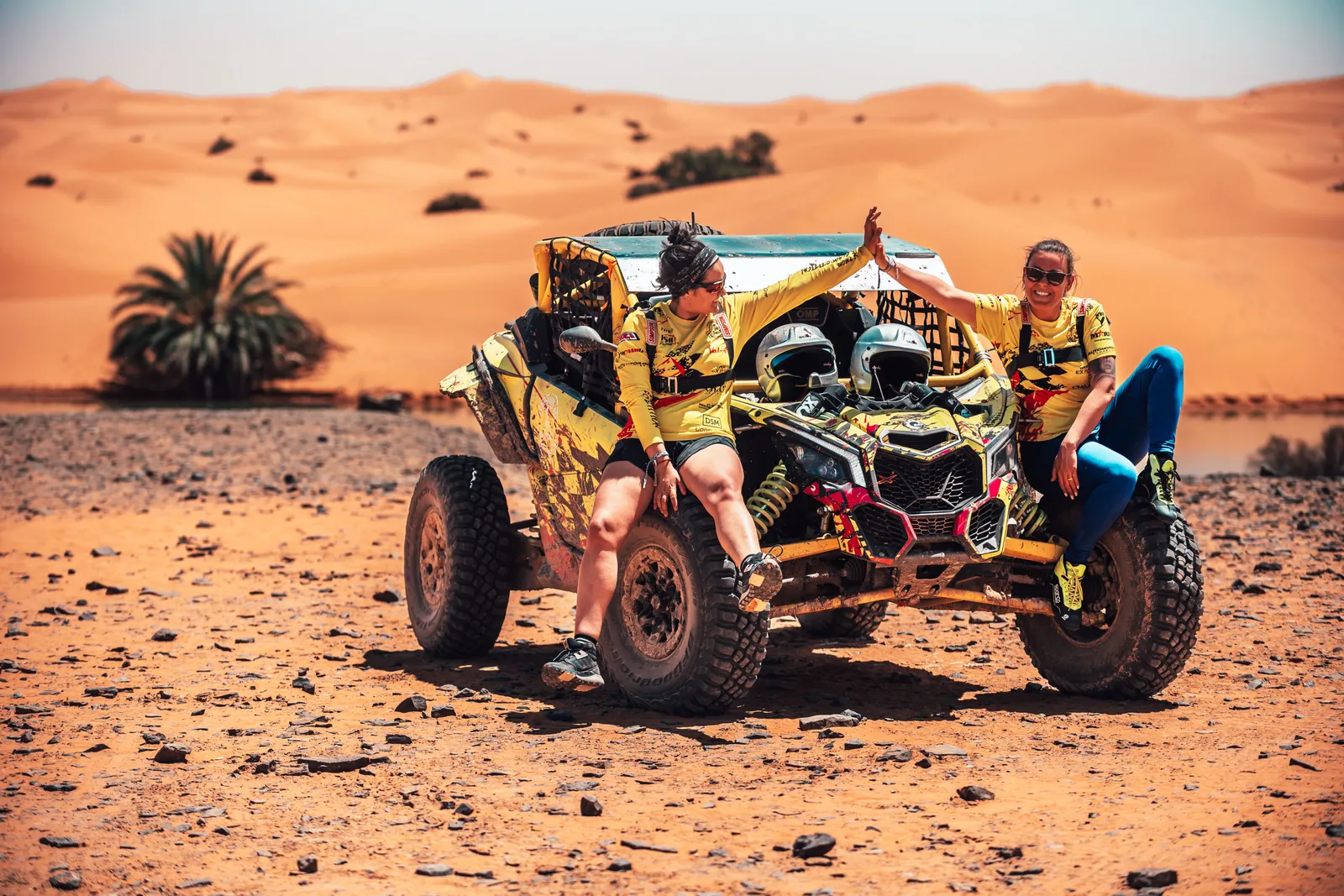 Dos participantes de rally chocando la mano sobre un buggy Can-Am en las dunas de Marruecos