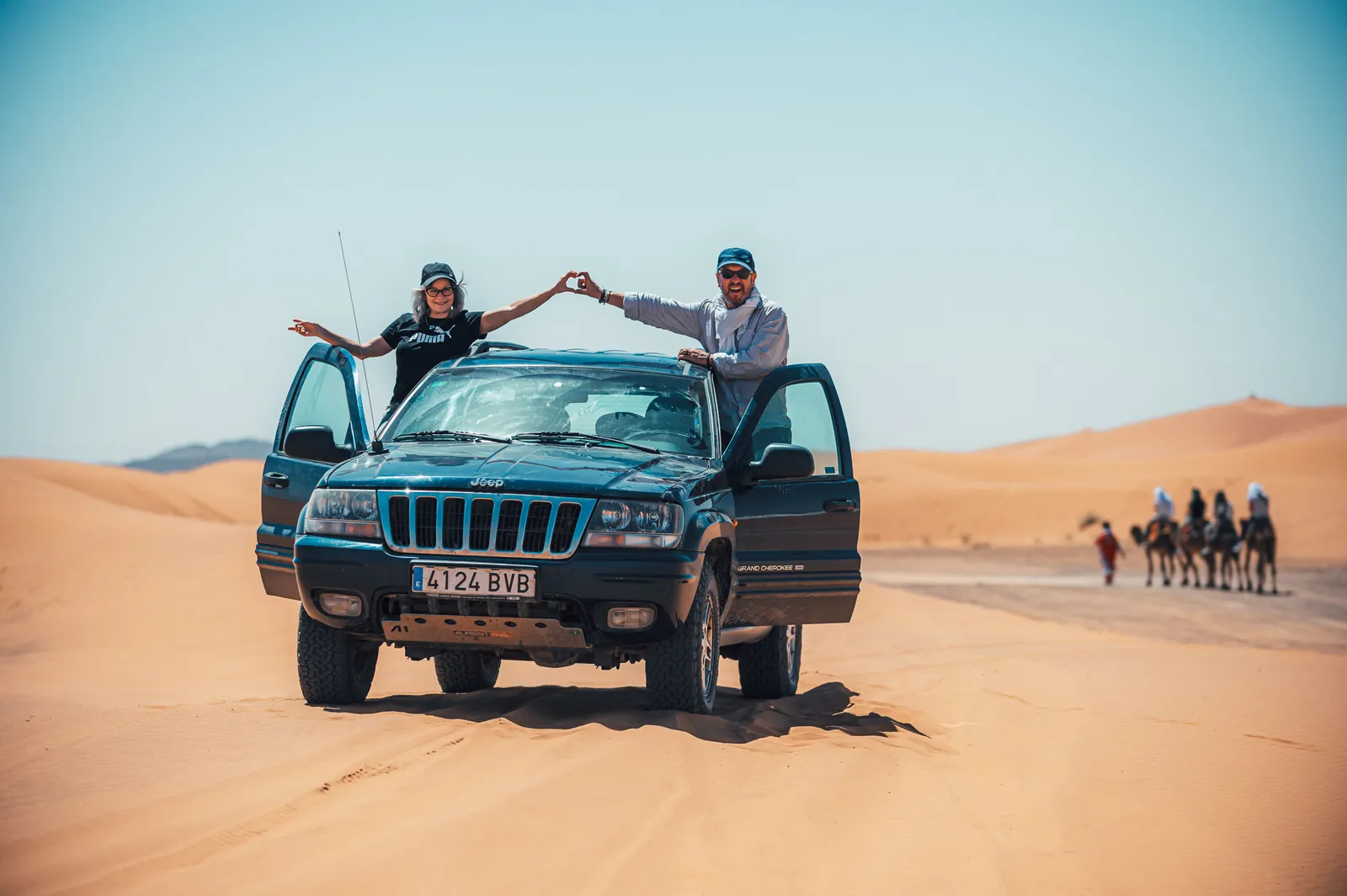 Pareja haciendo un corazón junto a su Jeep con una caravana de camellos de fondo en Marruecos